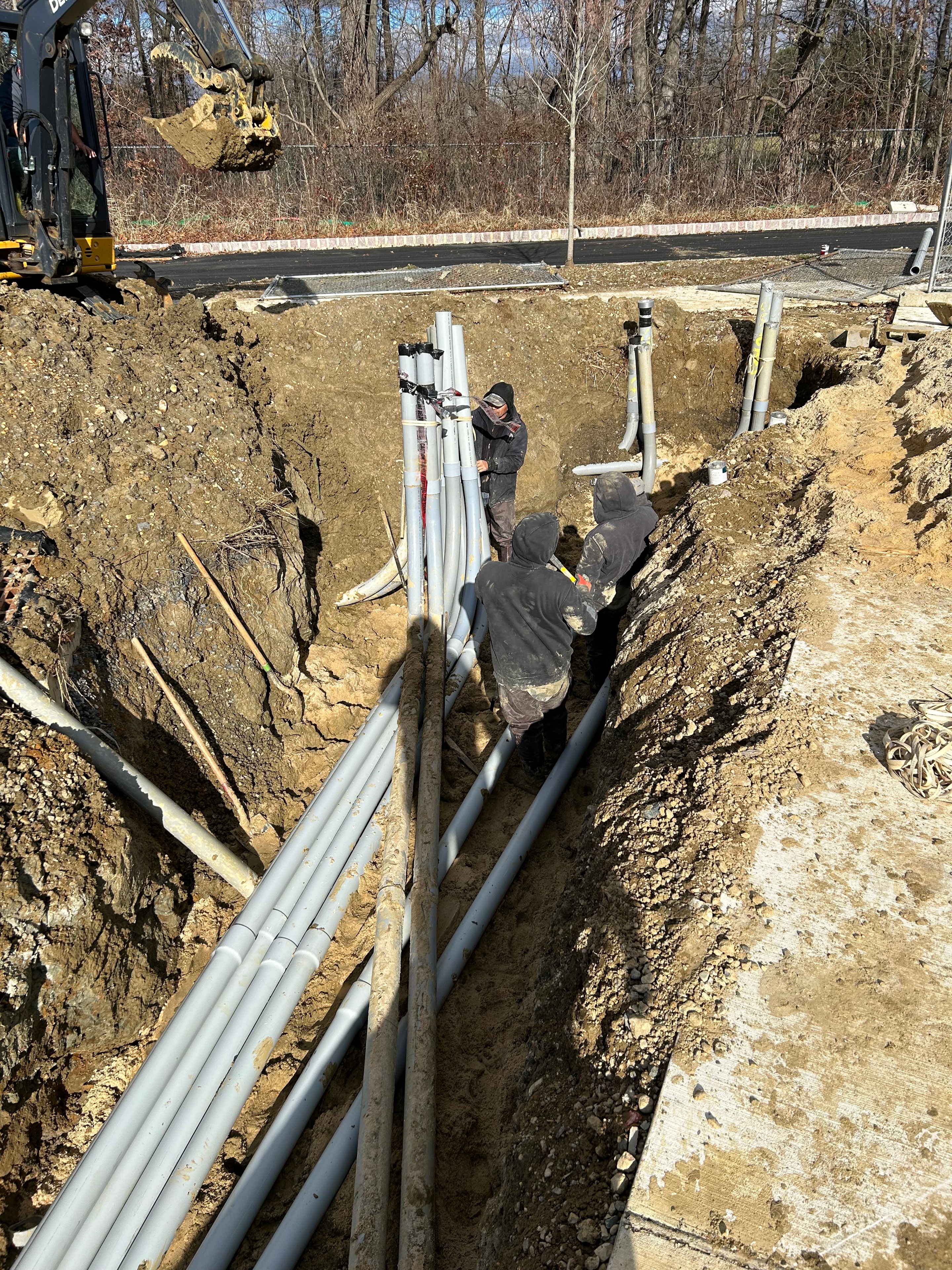 Two electricians installing multiple PVC conduits in deep excavated trench alongside road