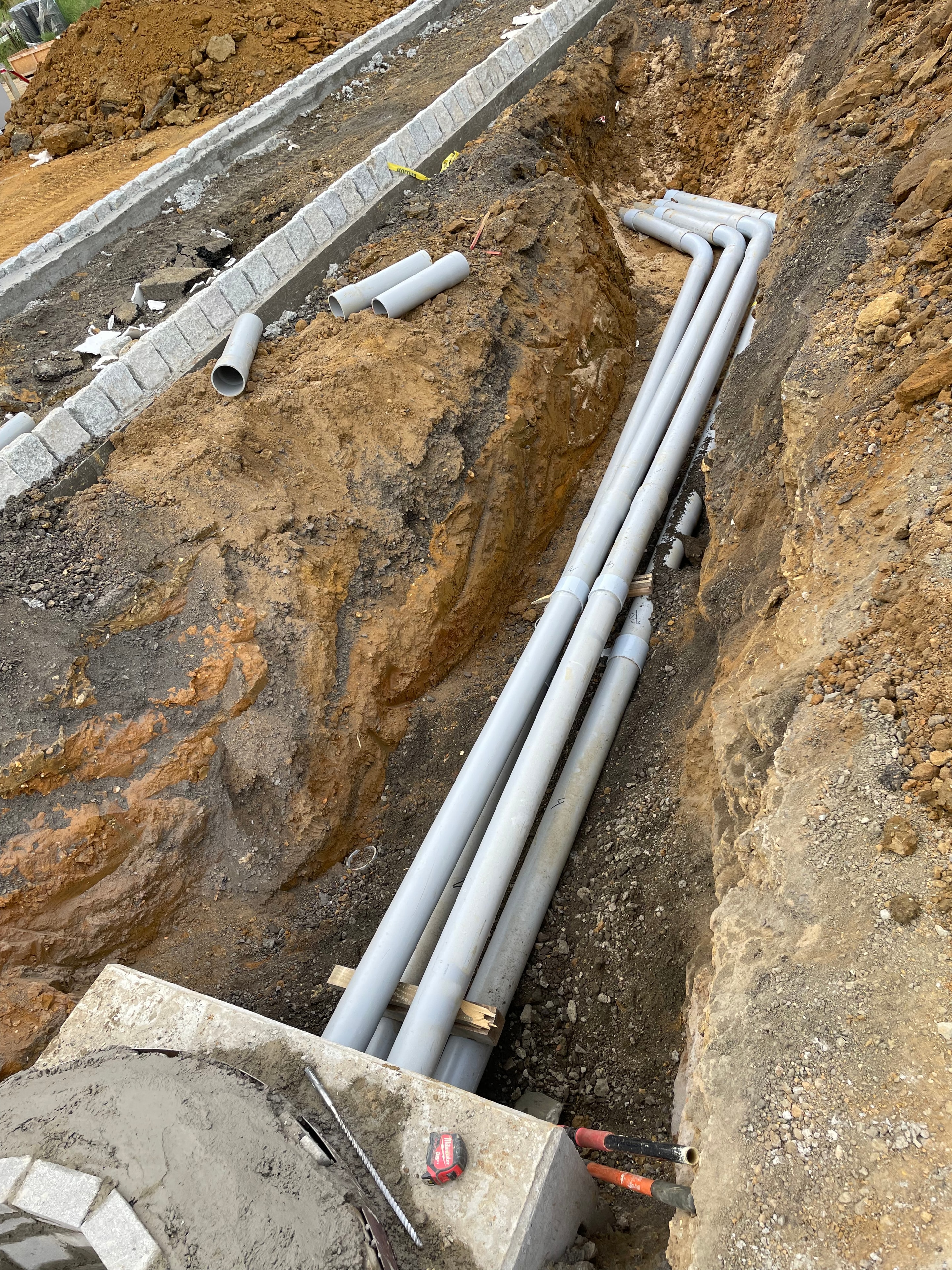 PVC conduit sweeps entering concrete manhole structure with Milwaukee tape measure in excavated trench
