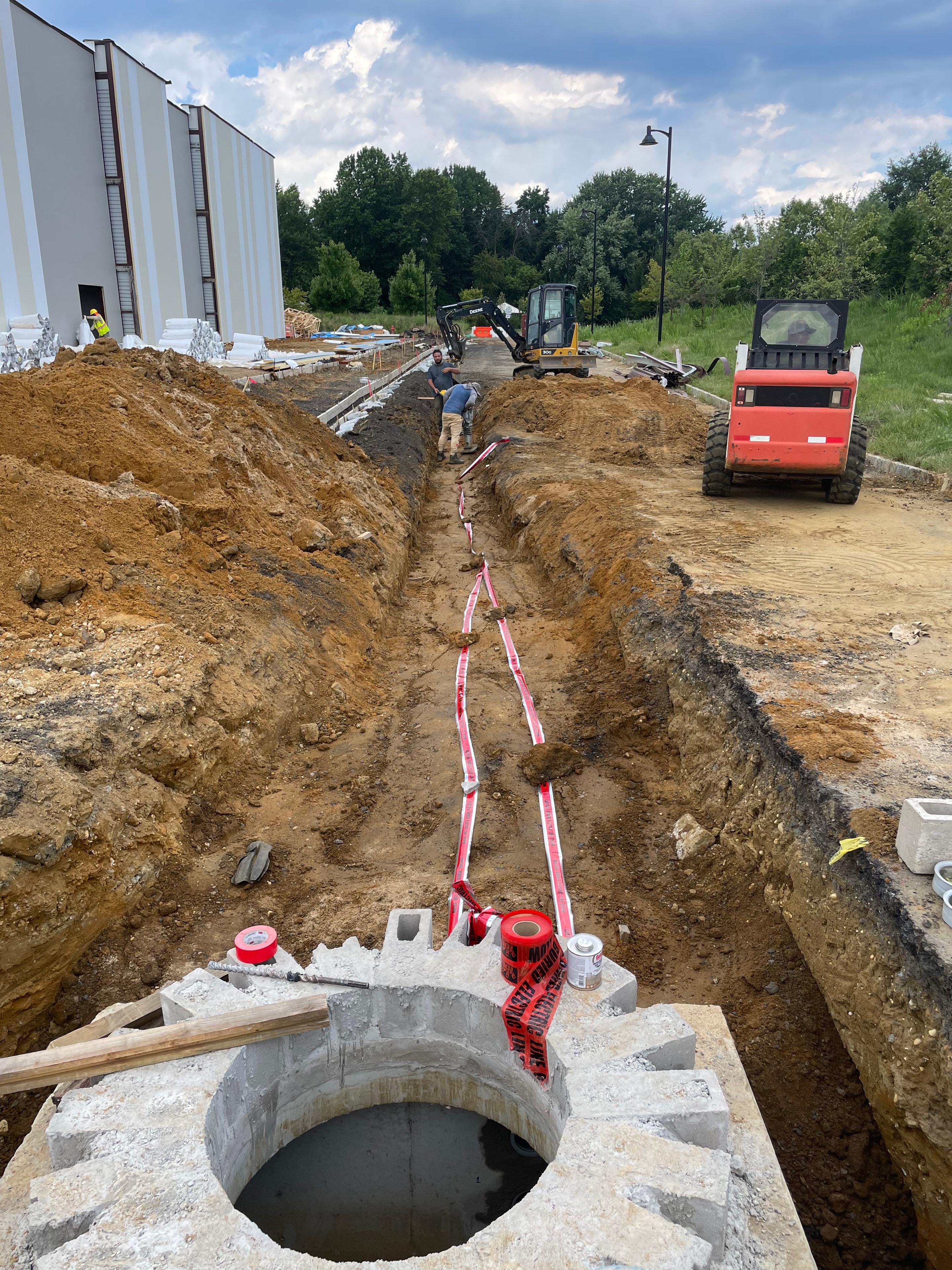 Open trench with concrete manhole ring and "Caution Buried Electric" tape at utility primary conduit installation
