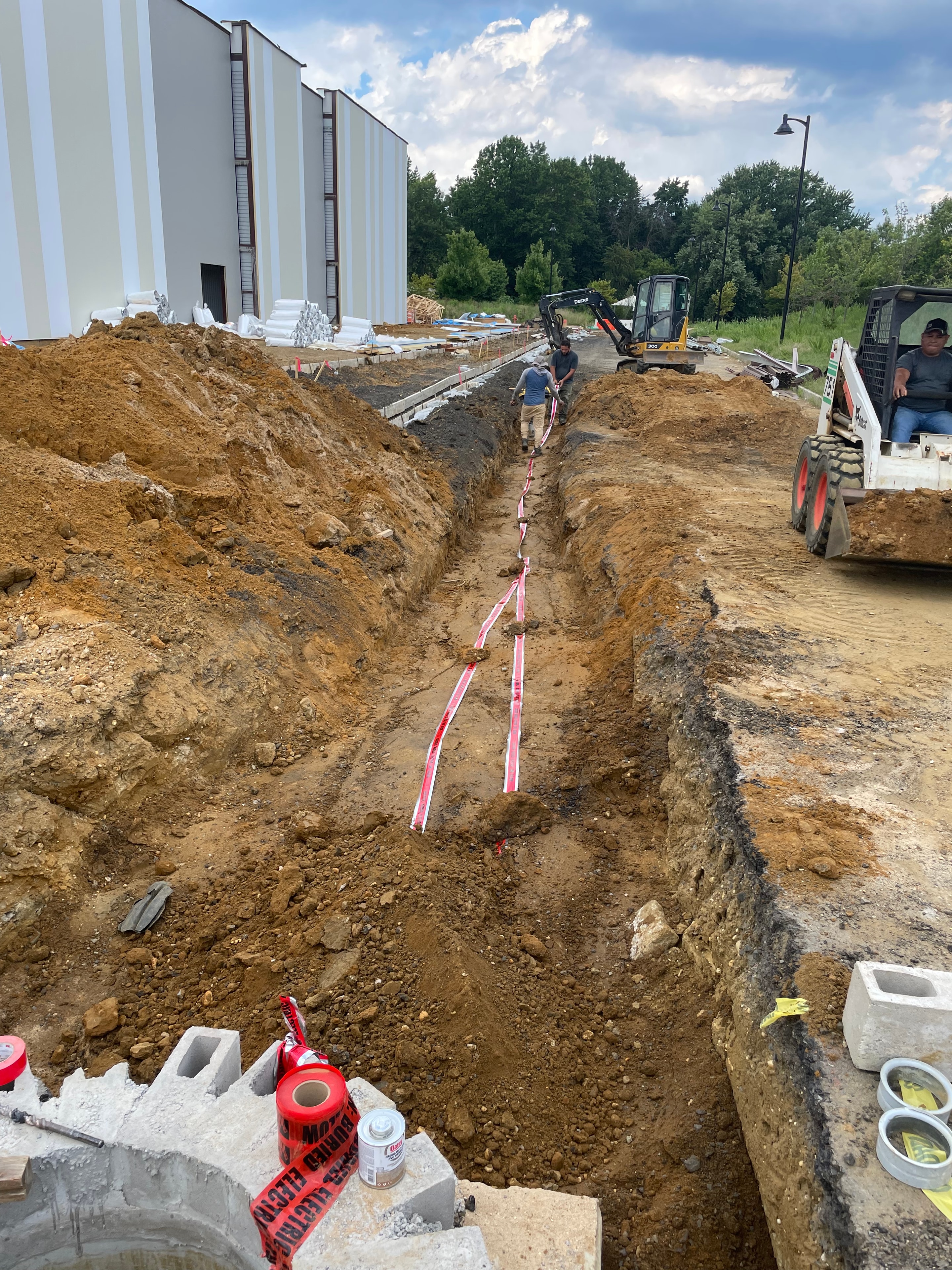 Wide excavation with "Buried Electric" warning tape and mini excavator at utility primary conduit site next to apartment building