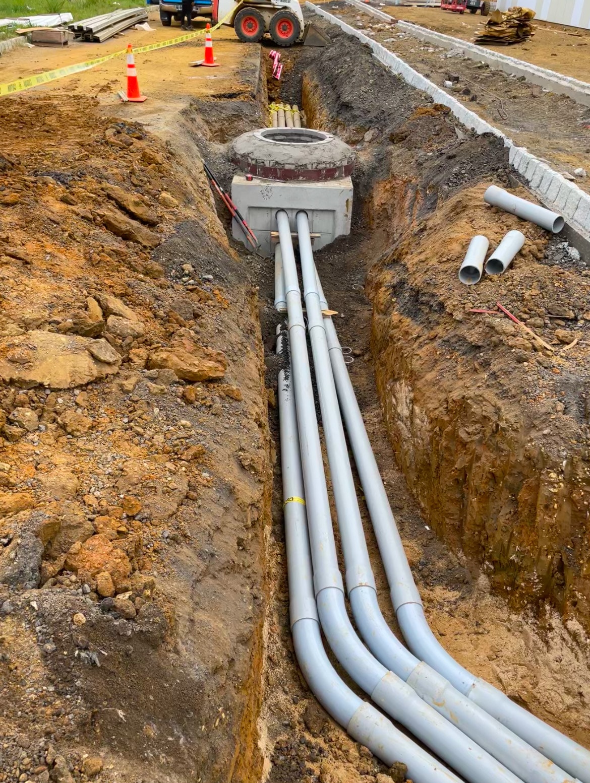 Four PVC primary conduits running through open trench to concrete manhole with Bobcat skid steer in background