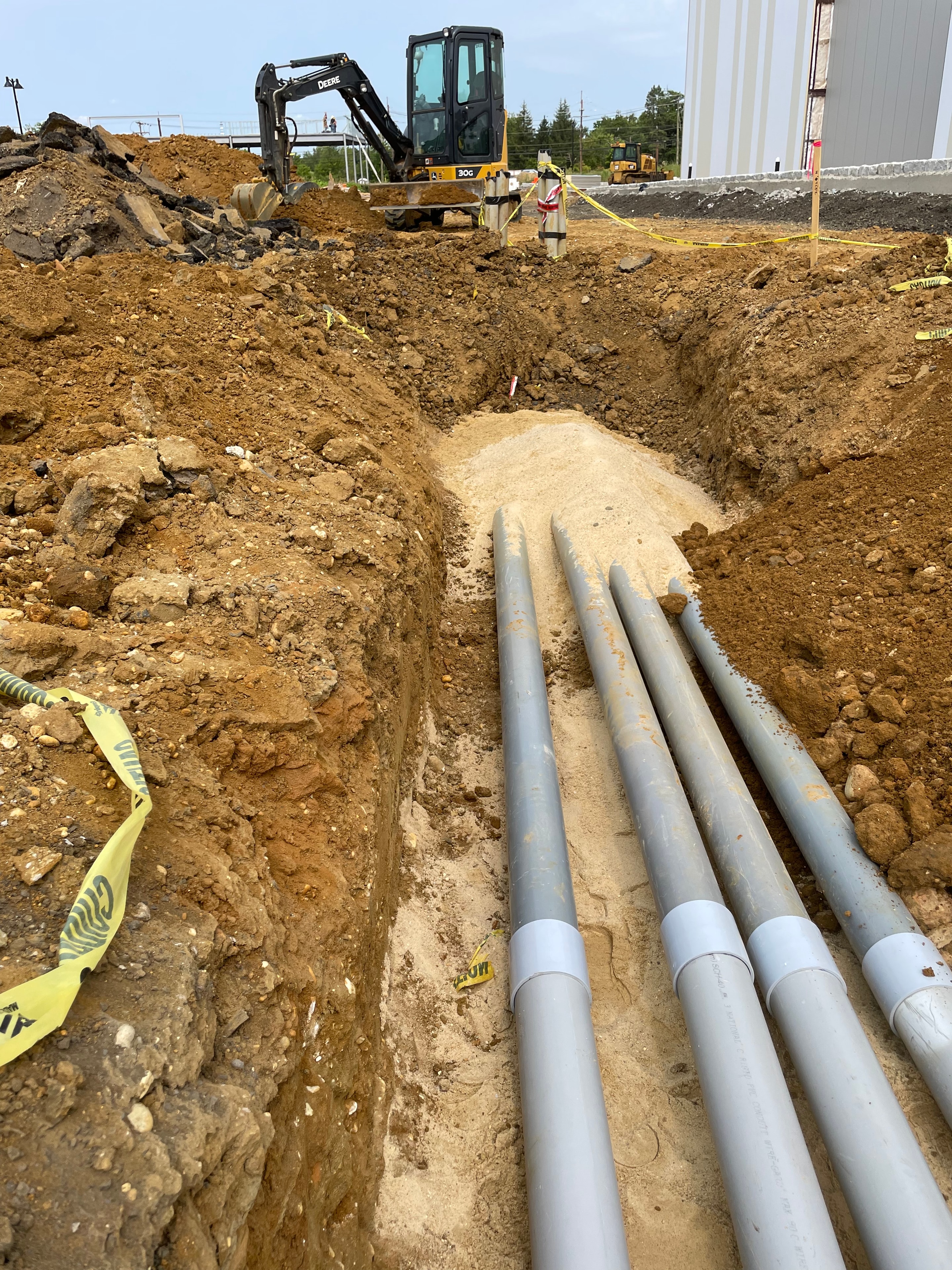 Five PVC primary conduits fanning out from trench with John Deere 30G excavator and caution tape at active site