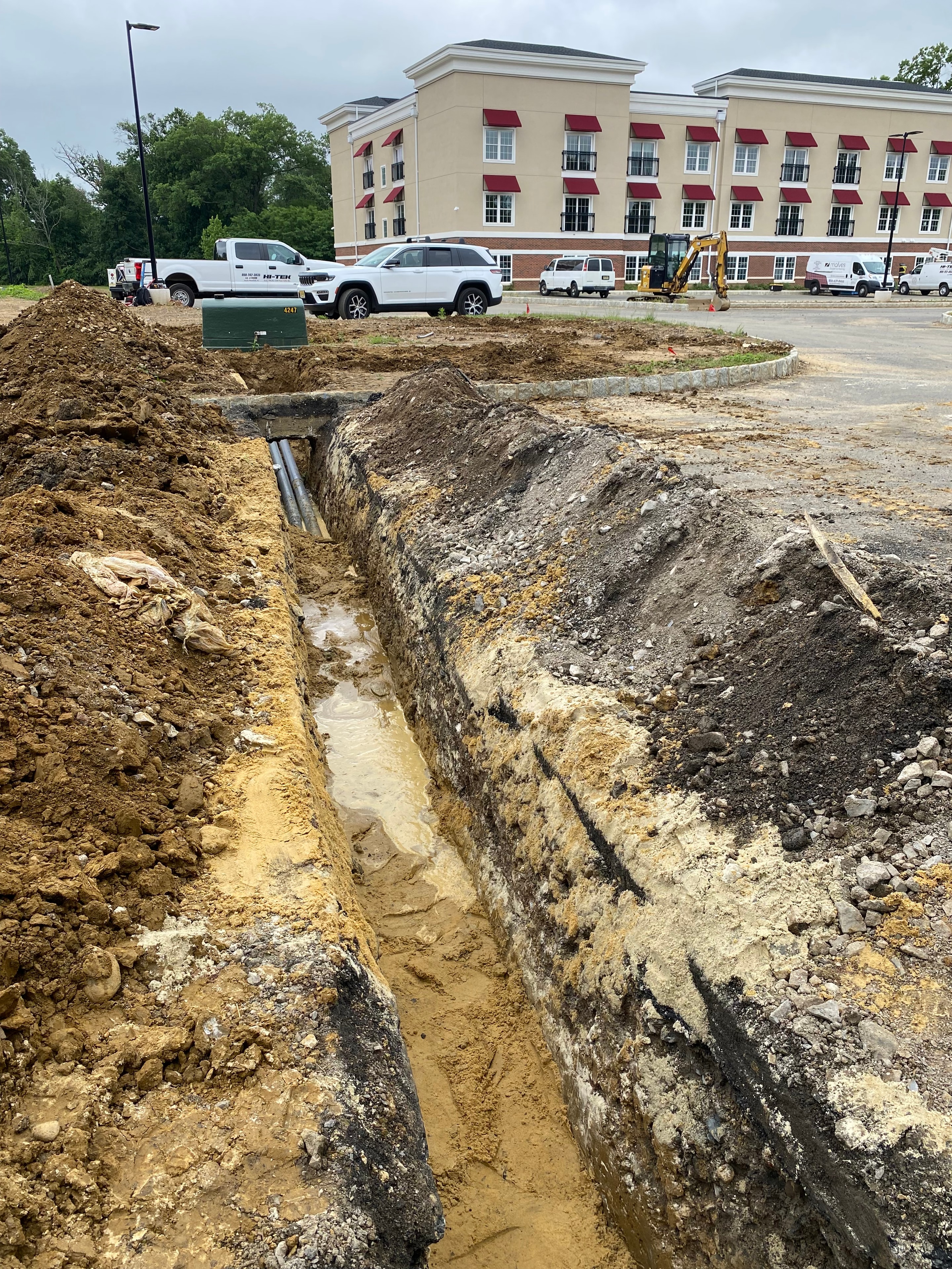 Deep excavation with conduit stubs visible at base of trench next to multi-story apartment building with Hi-Tek truck on site