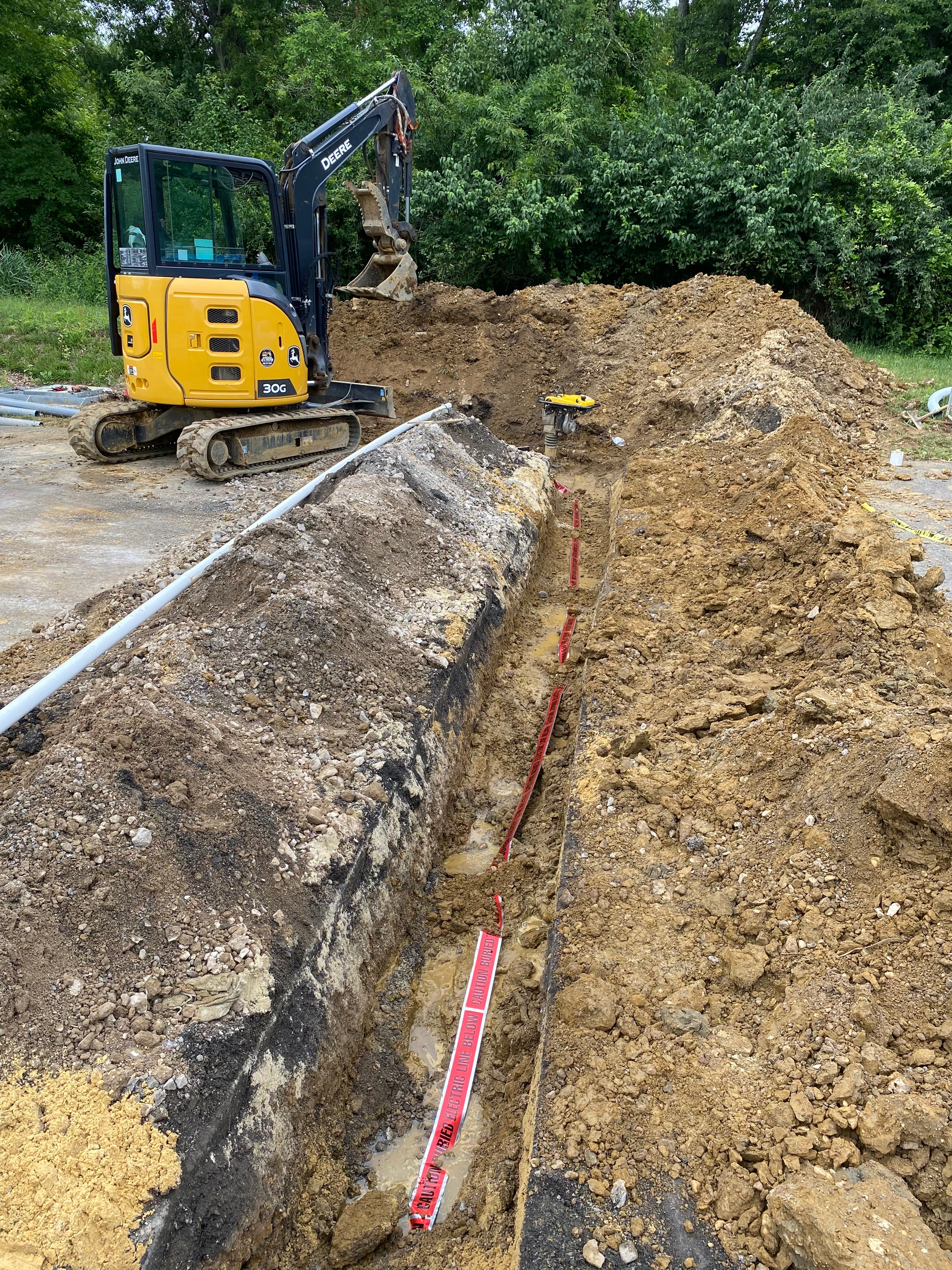 Crew installing galvanized steel conduits in wet rocky trench with John Deere excavator and Hi-Tek truck on site