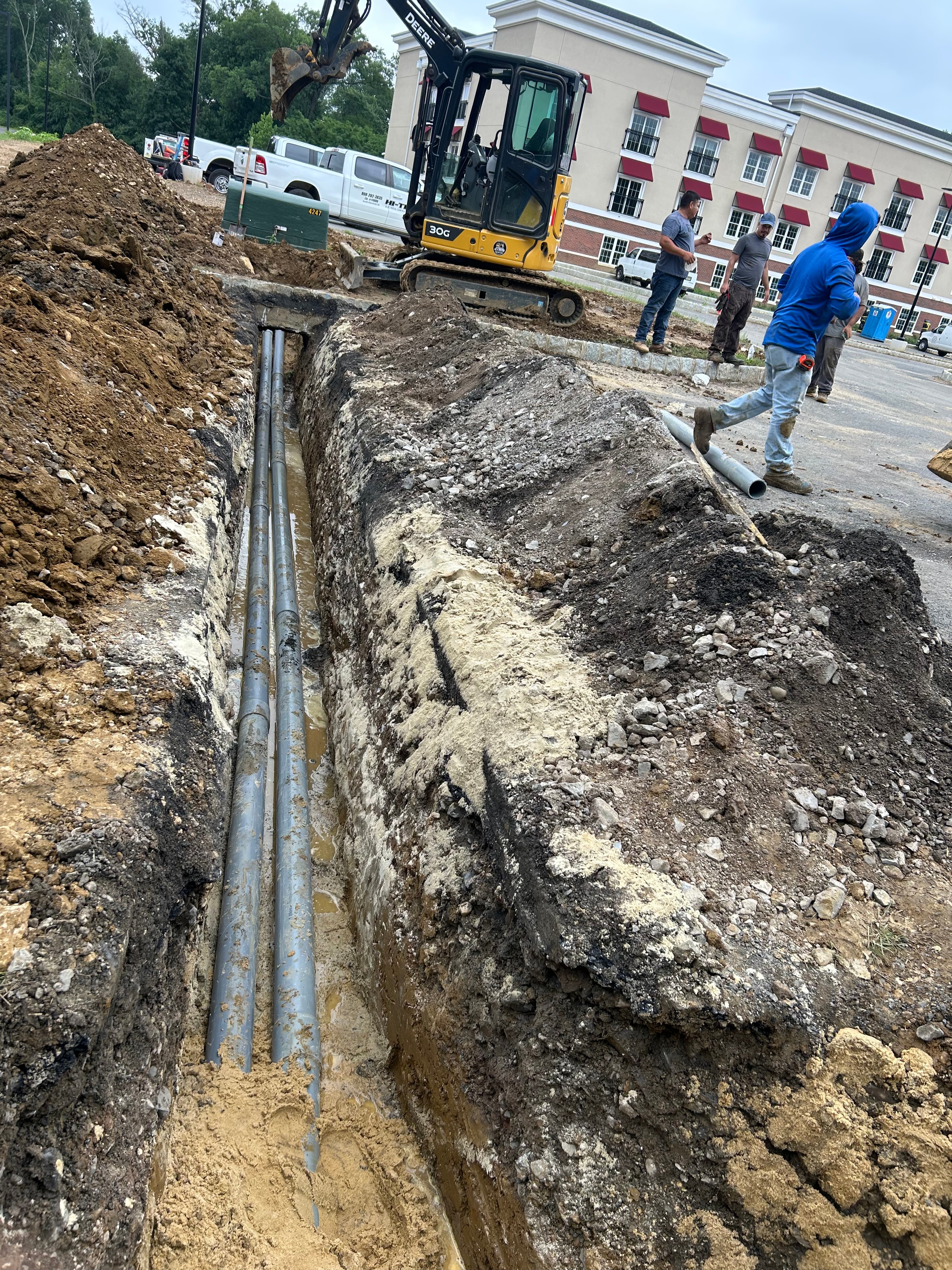 Aerial view of excavator bucket over trench with three PVC conduits laid beside green-tinted concrete pad
