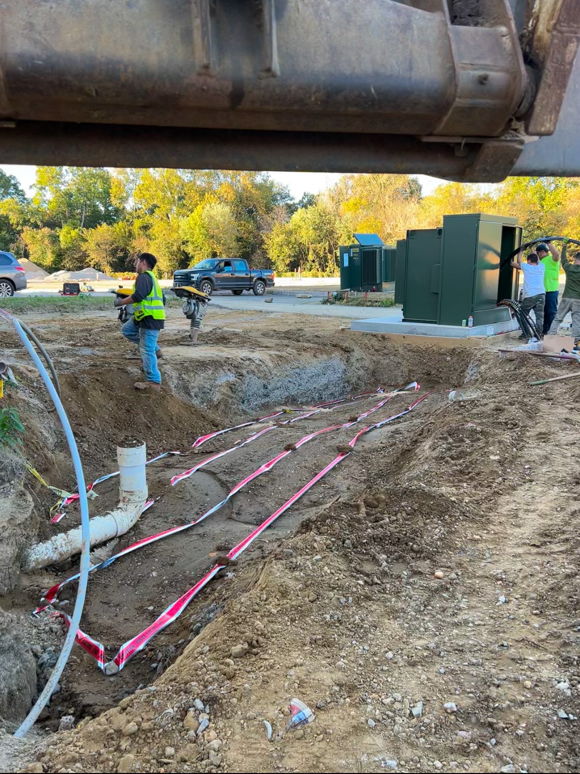 Crew working at utility transformer relocation site with green pad-mount transformer, buried electric warning tape, and PVC conduit stubs in open trench